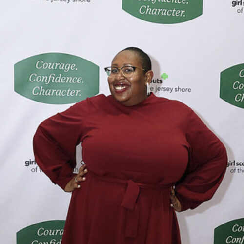 Dark skin woman wearing a short red dress with the girl scouts backdrop behind her.