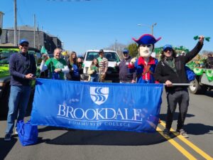Group of men and women and mascot standing behind a blue Brookdale banner in the street of the parade.