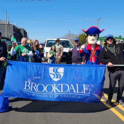 Group of men and women and mascot standing behind a blue Brookdale banner in the street of the parade.