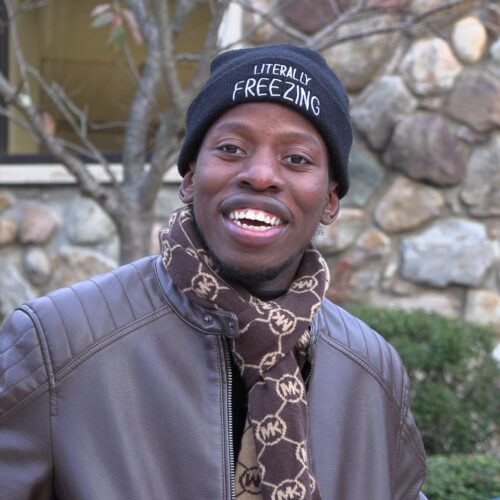 Dark skinned man with a hat on and scarf around his neck, smiling.