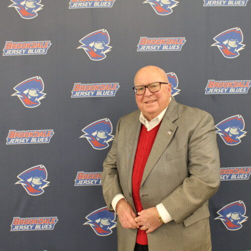 Dr. Lester Richens, wearing a sportscoat, red sweater and open collar shirt. He is standing in front of the Jersey Blues backdrop.