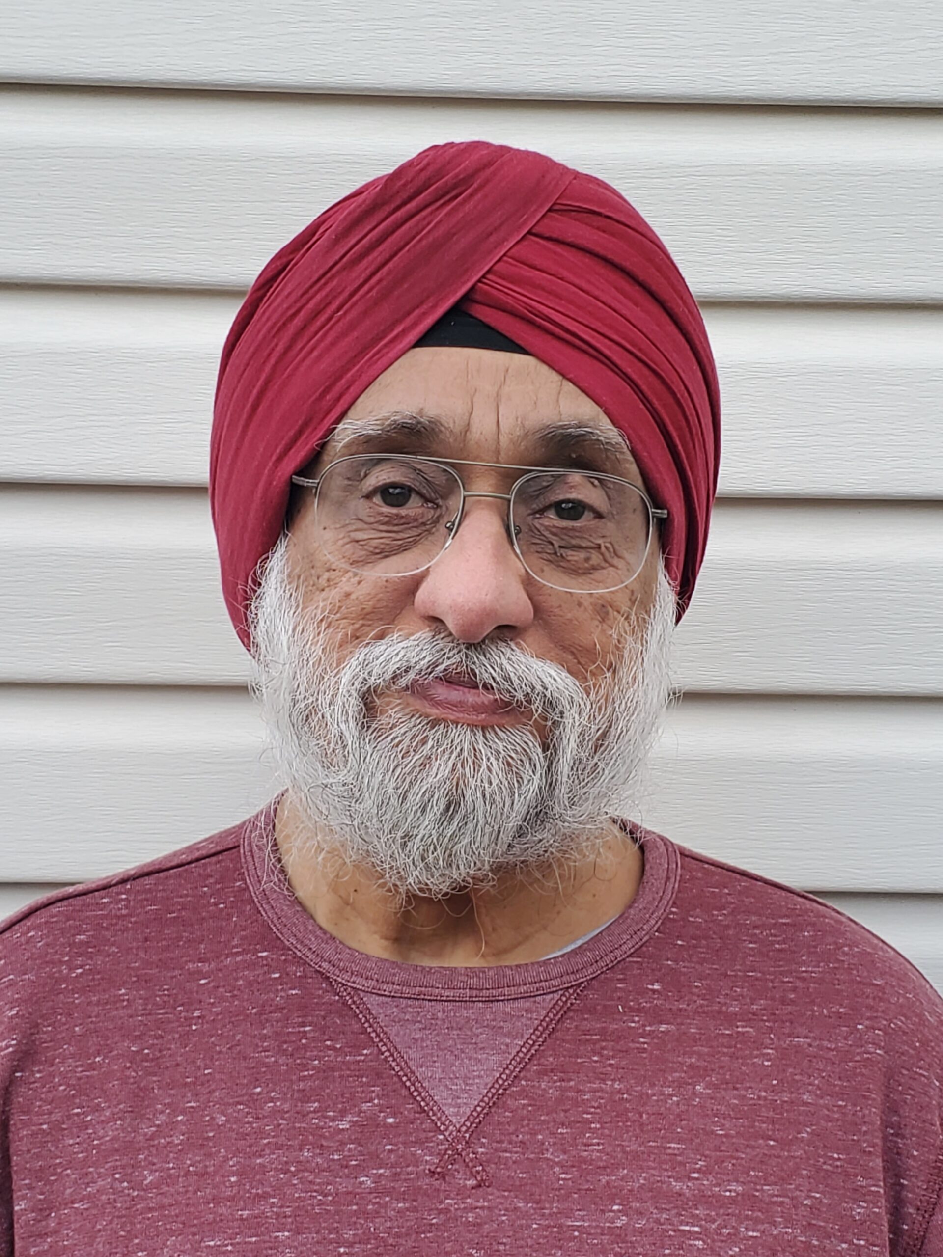 Man wearing a red turbin and red shirt. Has a grey beard and mustach and wears glasses.