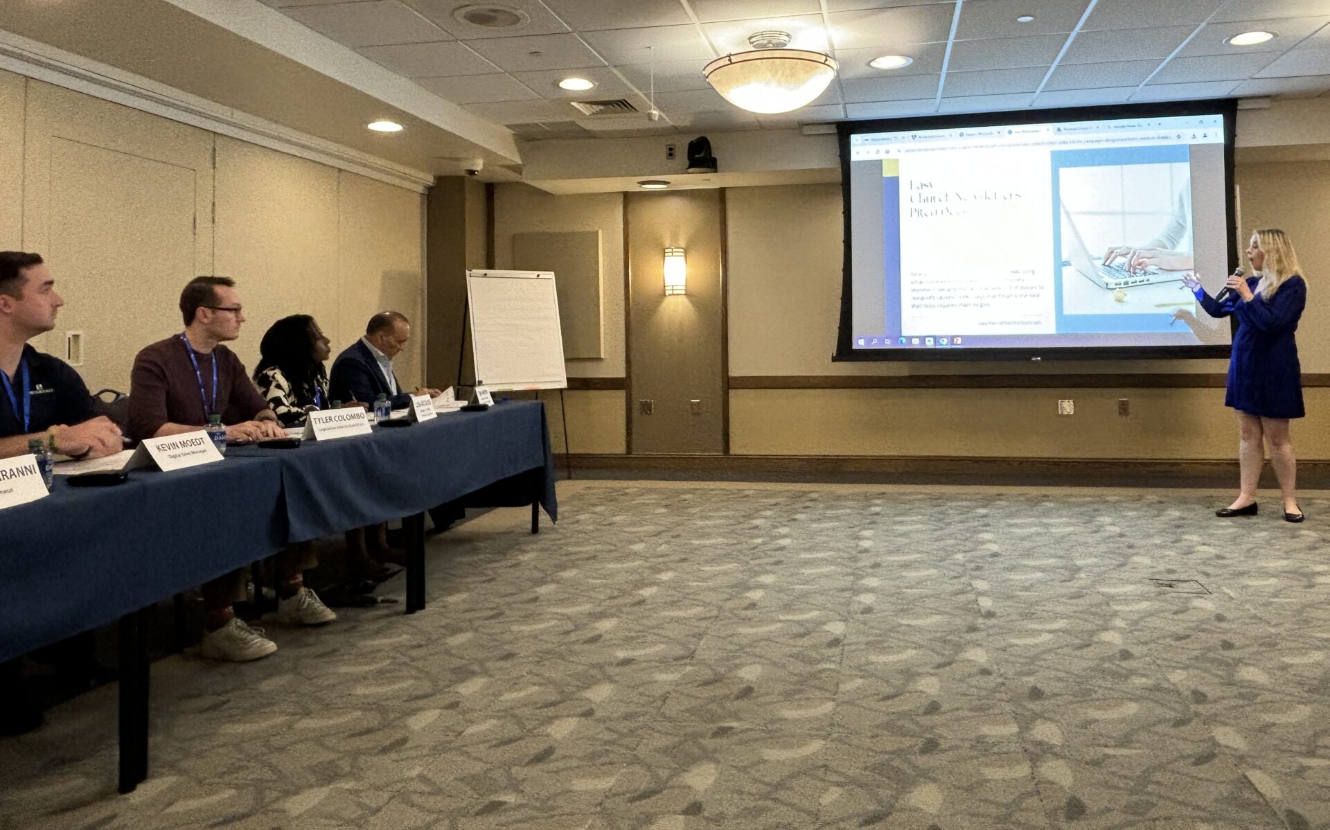 Woman presenting project using a screen with powerpoint of her pitch. Judges at table are listening to the presentation.