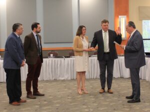Man wearing a suit and tie being sworn in by another man wearing a suit and tie with his wife wearing a white dress and tan sweater, holding a book that his hand is on. Two other men are looking on who are dressed in suit and ties.