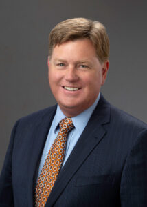 Man with blonde hair smiling, wearing a navy blazer, light blue shirt and gold tie.