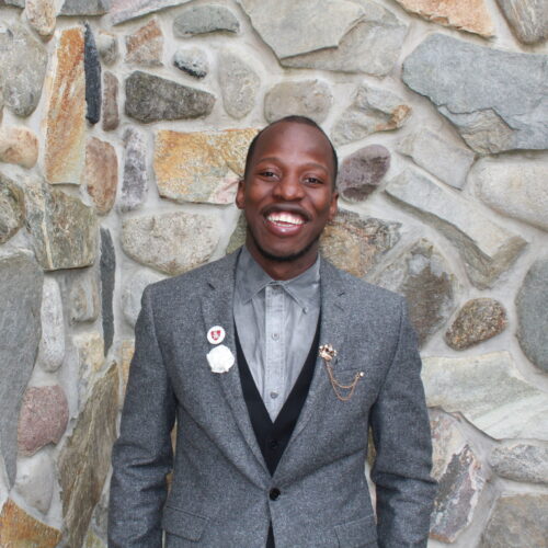 A man wearing a suit jacket, vest and collared shirt standing against a stone backdrop.
