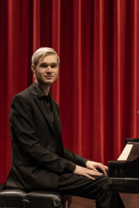 Man with blond hair and black shirt and jacket sitting at the piano.