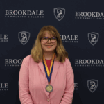 Woman with blond hair wearing glasses and a pink blazer and red shirt. Has a medal around her neck.