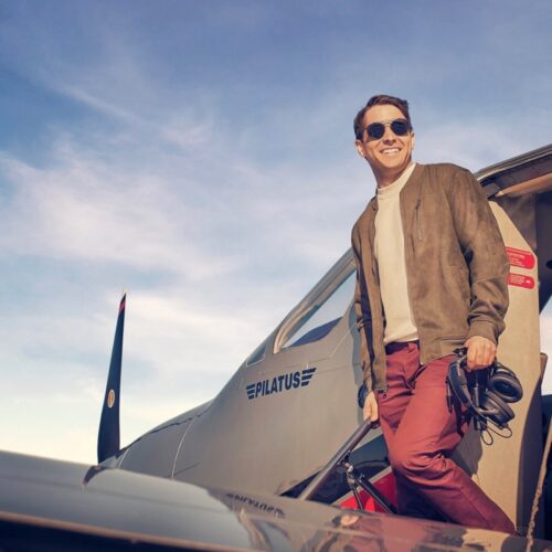 Mike Yuhas just outside an airplane. Wearing sunglasses, red pants, yellow top and brown jacket.