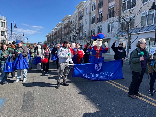 A group of people and the Jersey Blues mascot march in a St. Patrick’s Day parade, carrying a blue “Brookdale Community College” banner while wearing casual clothing with green accents along a city street lined with spectators and apartment buildings.