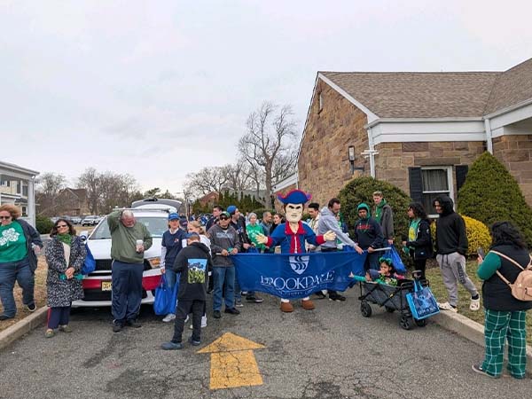 Adults and children gather with the Jersey Blues mascot on a residential street for a St. Patrick’s Day parade, holding a blue “Brookdale Community College” banner while wearing casual clothing with green accents near parked vehicles and townhouses.