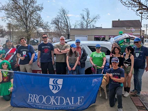 A group of adults and children pose together at a St. Patrick’s Day parade holding a blue “Brookdale Community College” banner, with participants wearing casual clothing and green accents along a neighborhood street.