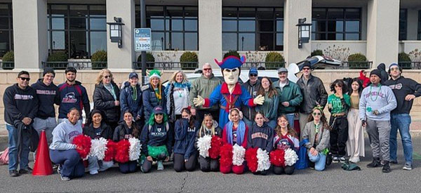 A large group poses for a group photo with the Jersey Blues mascot and red-and-white pom-poms outside a building during a St. Patrick’s Day celebration.